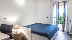 A single bed with a blue bedspread, a small table with a lamp, a wicker side table, and a view of the outdoors through a door. - N256 - Numana, brand new three-room apartment with garden (Sirolo)
