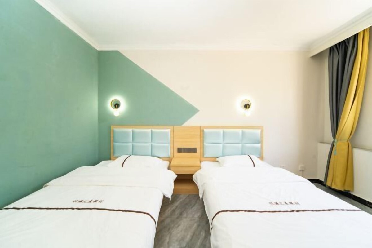 A hotel room with two beds, a light-colored wall, a wooden headboard, and a curtain on the right.