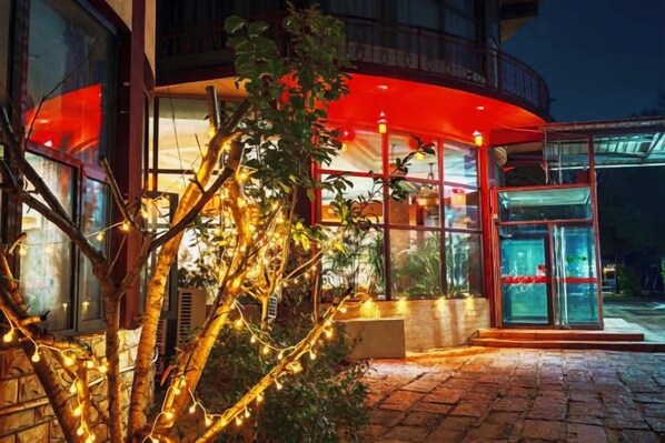 Entrada de hotel con puerta de vidrio, toldo rojo y un árbol con luces.