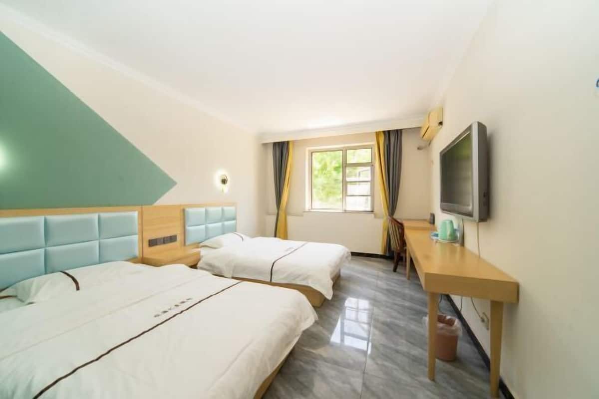 Ein Hotelzimmer mit zwei Betten, einem Fernseher, einem Schreibtisch und einem fensterbelaubten Fenster.