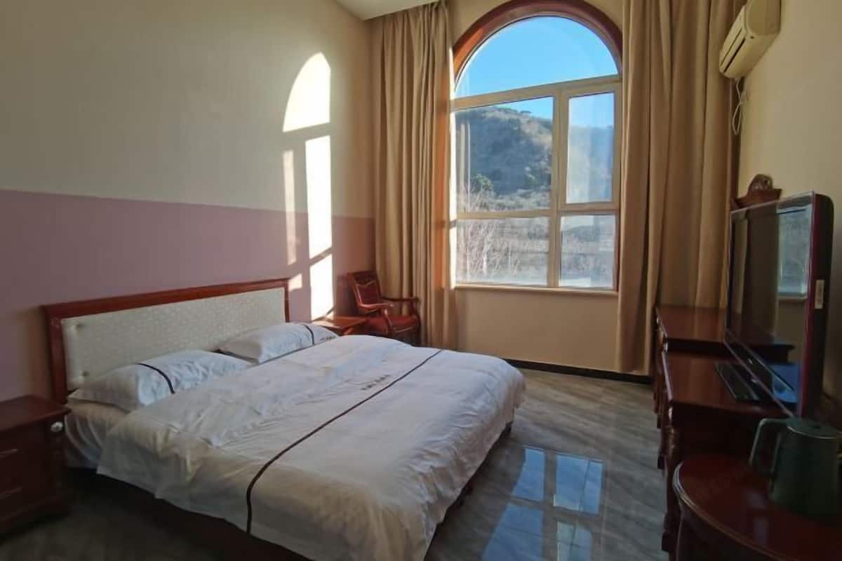 Ein Hotelzimmer mit Bett, Schreibtisch, Stuhl, einem großen Fenster mit Vorhängen und einem Fernseher.
