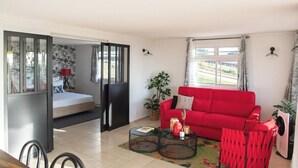 A living room with a red sofa, a dining table with chairs, a small coffee table with decorative items, a sliding door leading to a bedroom, and a large window with curtains.