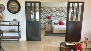 A room with a bed, a desk, and a chair. There is a door leading to another room with a bed and a nightstand.