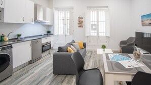 Una cucina moderna con mobili bianchi, forno integrato e lavastoviglie. È presente un divano grigio con cuscini e un tavolo da pranzo con sedie.