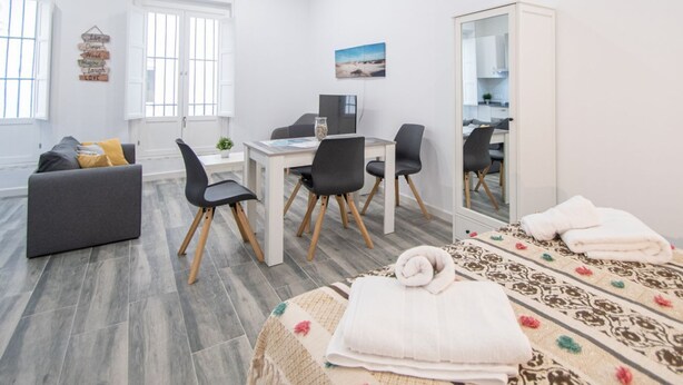 Una stanza moderna e luminosa con un divano grigio, un tavolo da pranzo con sedie, uno specchio grande e un letto con copripiumino decorato.
