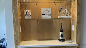 A wooden cabinet with a glass shelf holding wine glasses, a welcome sign, and decorative items. - Lake Winnipesaukee Townhouse (Gilford)