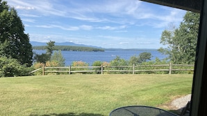 A view from a room with a metal table and a chair, overlooking a grassy area with a wooden fence and a body of water. - Lake Winnipesaukee Townhouse (Gilford)