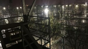 Vista noturna de uma varanda com guarda-corpo de metal, com vista para um estacionamento com vegetação escassa e postes de luz.