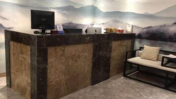 A reception desk with a computer monitor, a phone, and a small table with a vase of flowers. There is a bench with a cushion in the foreground.