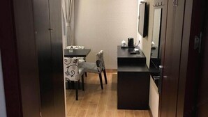 Um corredor estreito que dá para uma sala de jantar com mesa e cadeiras, uma pequena área de cozinha com bancada e eletrodomésticos, e uma televisão fixada na parede.