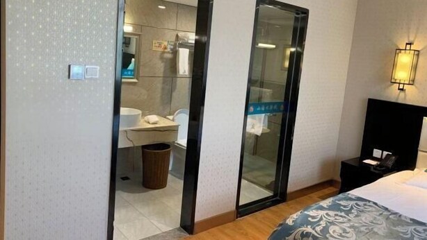 Quarto de hotel com cama, mesa de cabeceira, espelho, luminária na parede e banheiro visível através de uma porta de vidro.