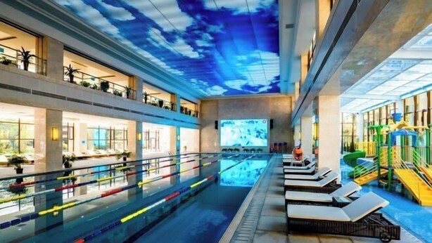 Une piscine intérieure dotée d’un plafond qui simule un ciel nuageux, entourée d’espaces de détente et d’un espace de jeux.