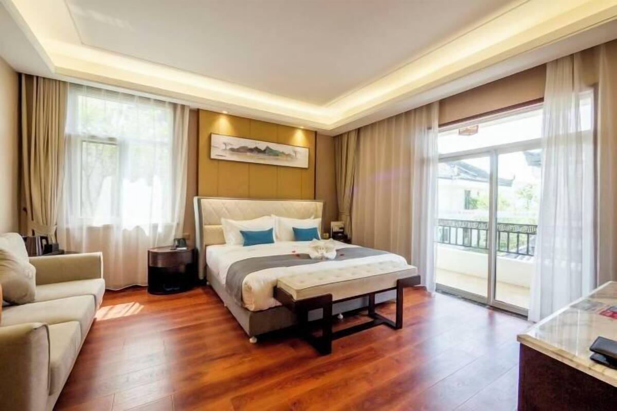 A hotel room with a bed, a sofa, a bedside table, a lamp, a window with curtains, and a balcony.