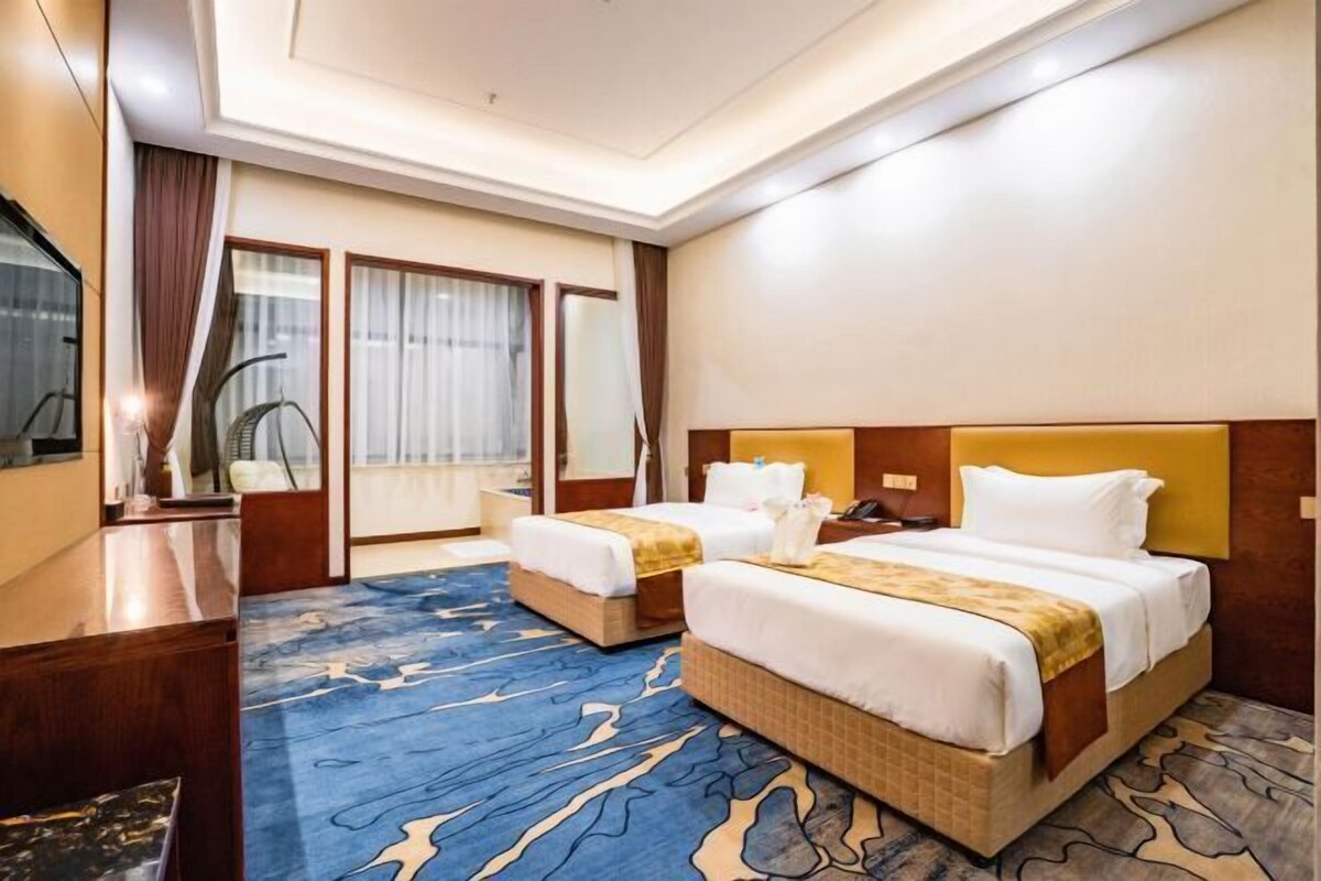 A hotel room with two beds, a desk, a chair, a large window with curtains, and a blue patterned carpet.