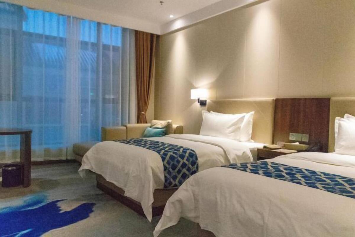 A hotel room with two beds, a nightstand, a lamp, and a view of the city through large windows.