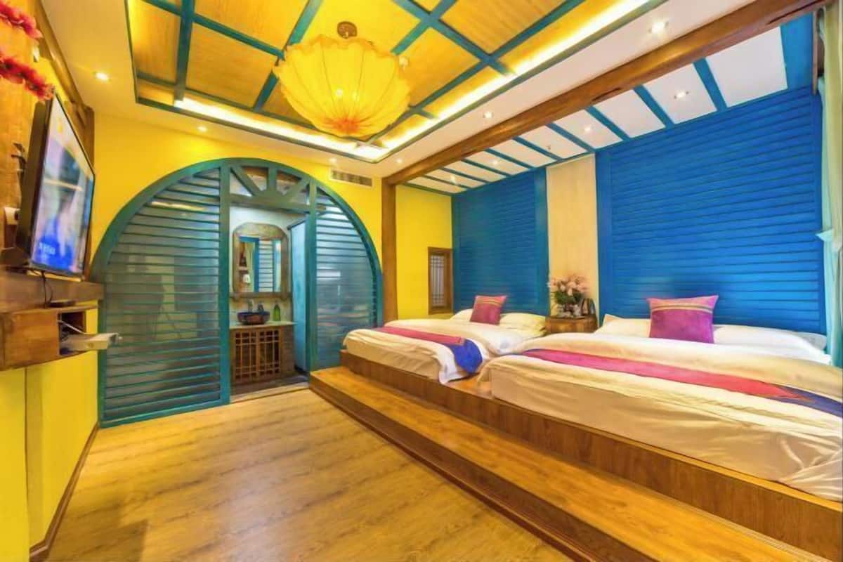 A hotel room with a wooden floor, two beds with colorful bedding, a large window with blue shutters, and a unique arched doorway.