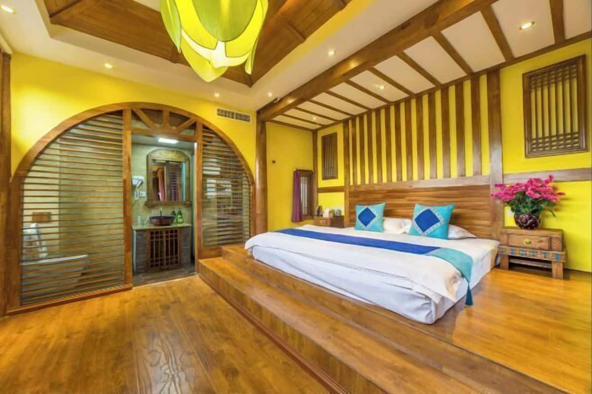 A bedroom with a large bed, wooden beams, a curved wooden door, and a small bedside table with a vase of flowers.
