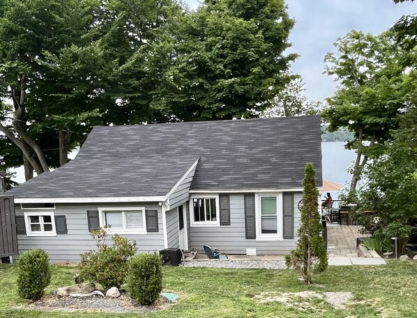 Une maison à deux étages, avec un toit gris et des éléments décoratifs blancs, entourée d’arbres et d’une pelouse.