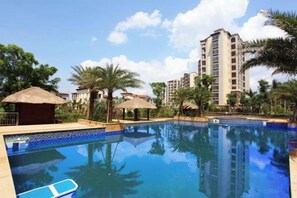 A swimming pool with a clear blue water surface, surrounded by palm trees and a thatched-roof structure. - Sincerity Holiday Apartment Hostel (Haikou)