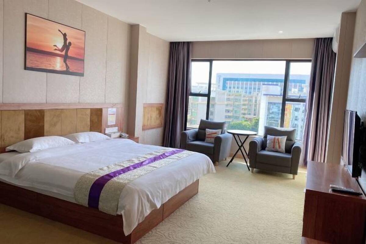 Ein Hotelzimmer mit einem großen Bett, zwei Sesseln, einem kleinen Tisch, einem Fernseher und Blick auf die Stadt.