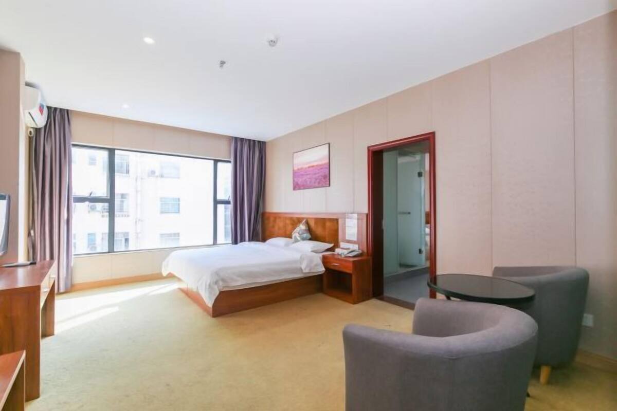 Ein Hotelzimmer mit einem Bett, einem Nachttisch, einer Lampe, zwei Sesseln, einem kleinen Tisch und einem Fenster mit Vorhängen.
