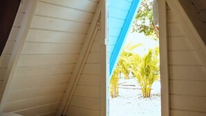 Ein Zimmer mit schrägem Dach und einem Fenster, das auf eine Sandfläche mit Palmen blickt.