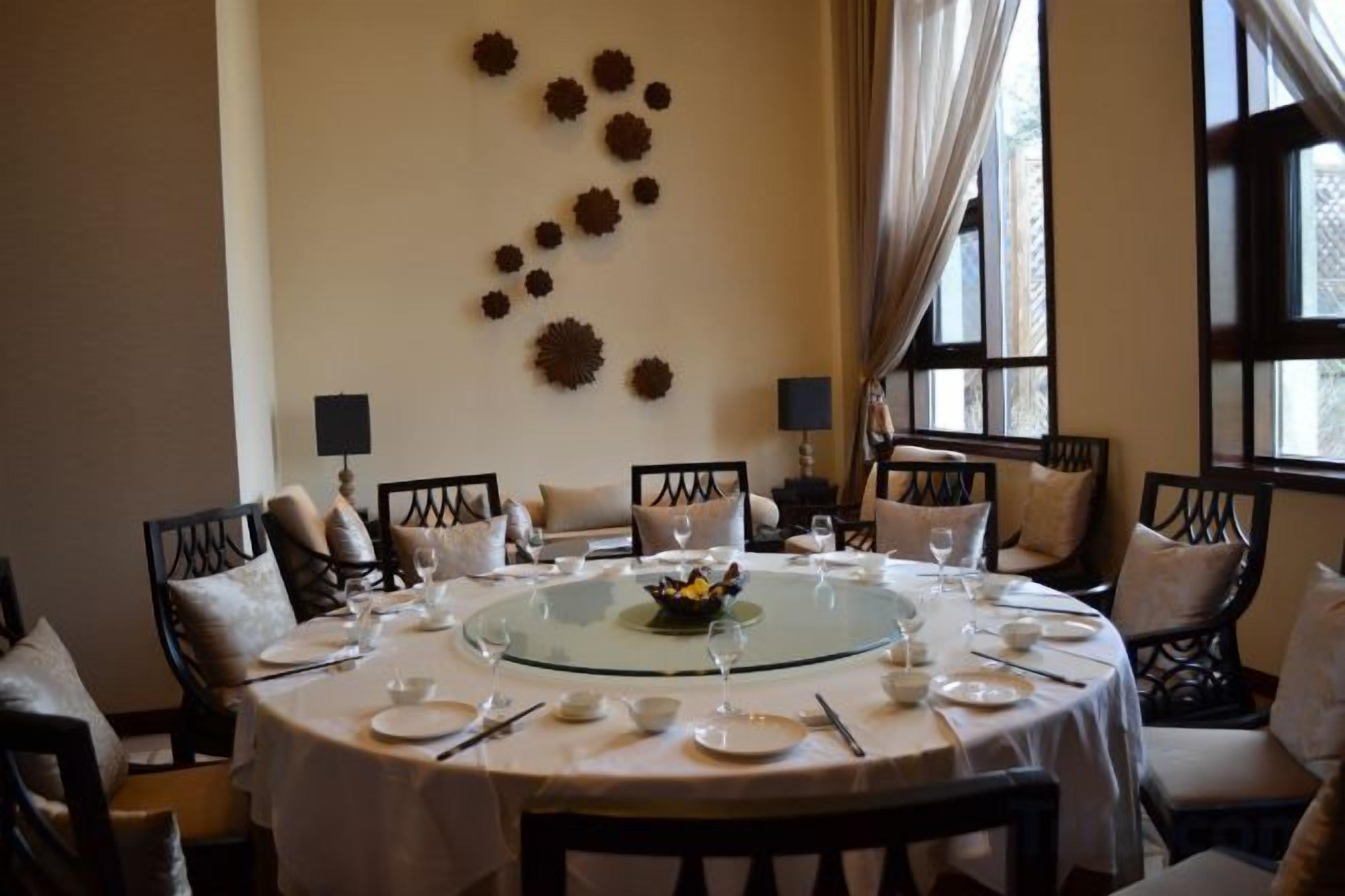 Un comedor con una mesa redonda dispuesta para una comida, rodeada de sillas, con un muro decorativo y ventanas al fondo.