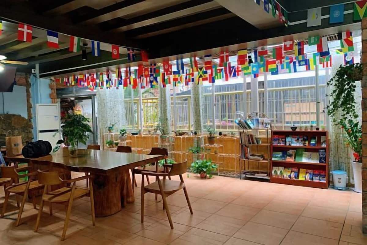 A library with a wooden table and chairs, surrounded by bookshelves and potted plants. Flags are displayed on the windows.