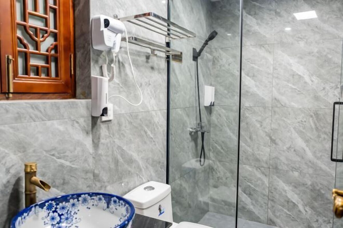 Un baño con pared de mármol, un inodoro, un lavabo con un cuenco azul con motivos florales, un secador de pelo en un soporte de pared y una ducha con puerta de vidrio.