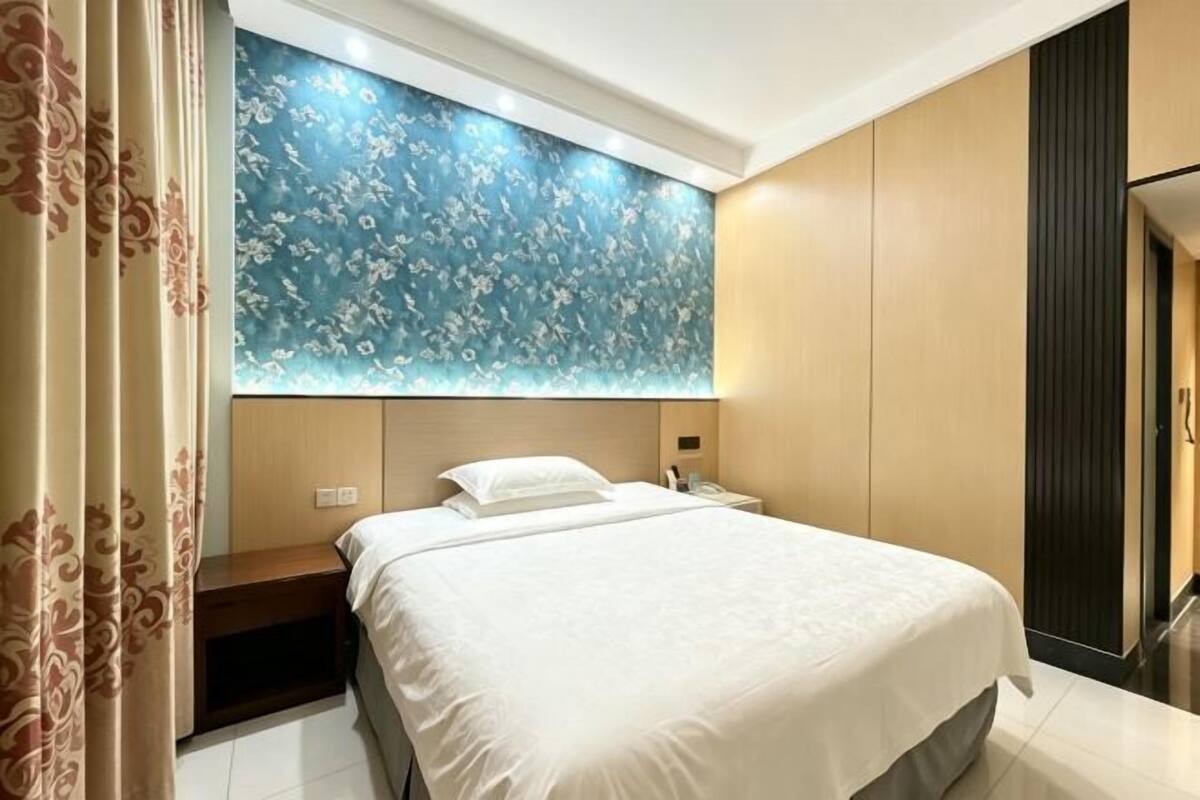 A modern hotel room with a large bed, a nightstand, a wall-mounted lamp, and a decorative wall panel.