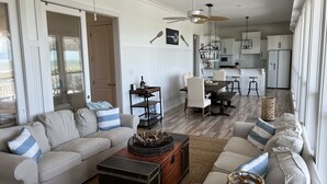 A spacious living area with a large sofa, a coffee table, and a dining table. There is a kitchen in the background with white cabinets and a refrigerator.