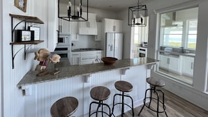 A modern kitchen with white cabinetry, granite countertops, and bar stools. There are pendant lights, a large window, and a view of the outdoors.