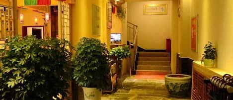 A hotel lobby with a stone floor, potted plants, a staircase, and a reception desk.