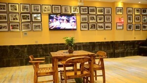 A dining area with a wooden table and chairs, a television mounted on the wall displaying a program, and a wall adorned with framed photographs. - Holiday Weekend Hotel (Xiahe)
