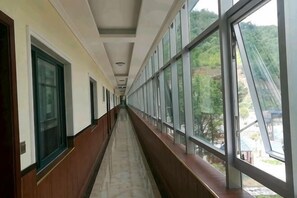 Un couloir doté de grandes fenêtres et de grandes portes, qui mène à un bâtiment offrant une vue sur les arbres et sur une structure bleue à l’extérieur.