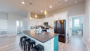 A spacious kitchen with white cabinets, a stainless steel refrigerator, and a granite countertop. There are three black bar stools and a window with blinds. - Private Pool and Golf Cart Included (Port Aransas)