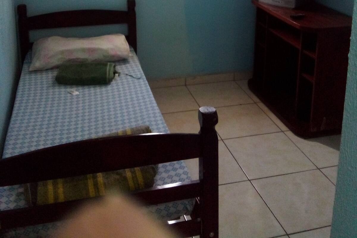 A small, single-bed room with a wooden headboard, a window with blinds, a standing fan, and a television on a wooden stand.
