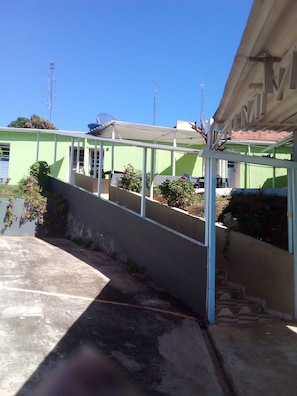 A covered outdoor area with a concrete floor, a metal railing, and a green building in the background. - Hotel pousada chic (Tatui)