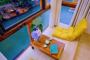 Una habitación con vista al río, una mesa de madera con un libro y una tetera, una silla amarilla y una planta.