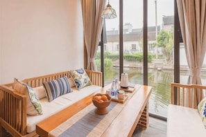 Una habitación con un sofá de madera, una mesa de comedor con un cuenco de fruta y vistas a un cuerpo de agua y edificios.