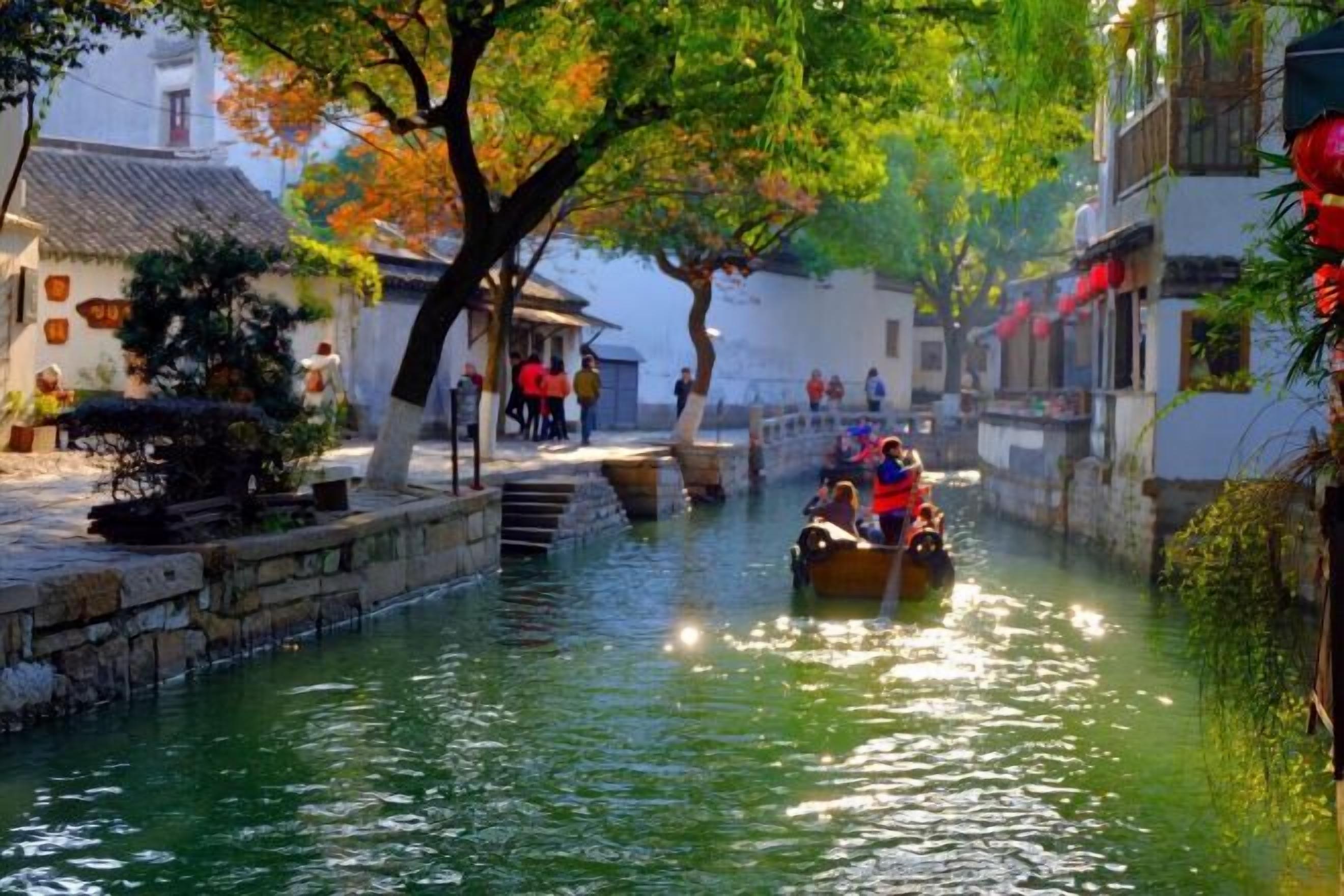 A narrow waterway with traditional buildings on either side, a boat with passengers, and a tree-lined path.