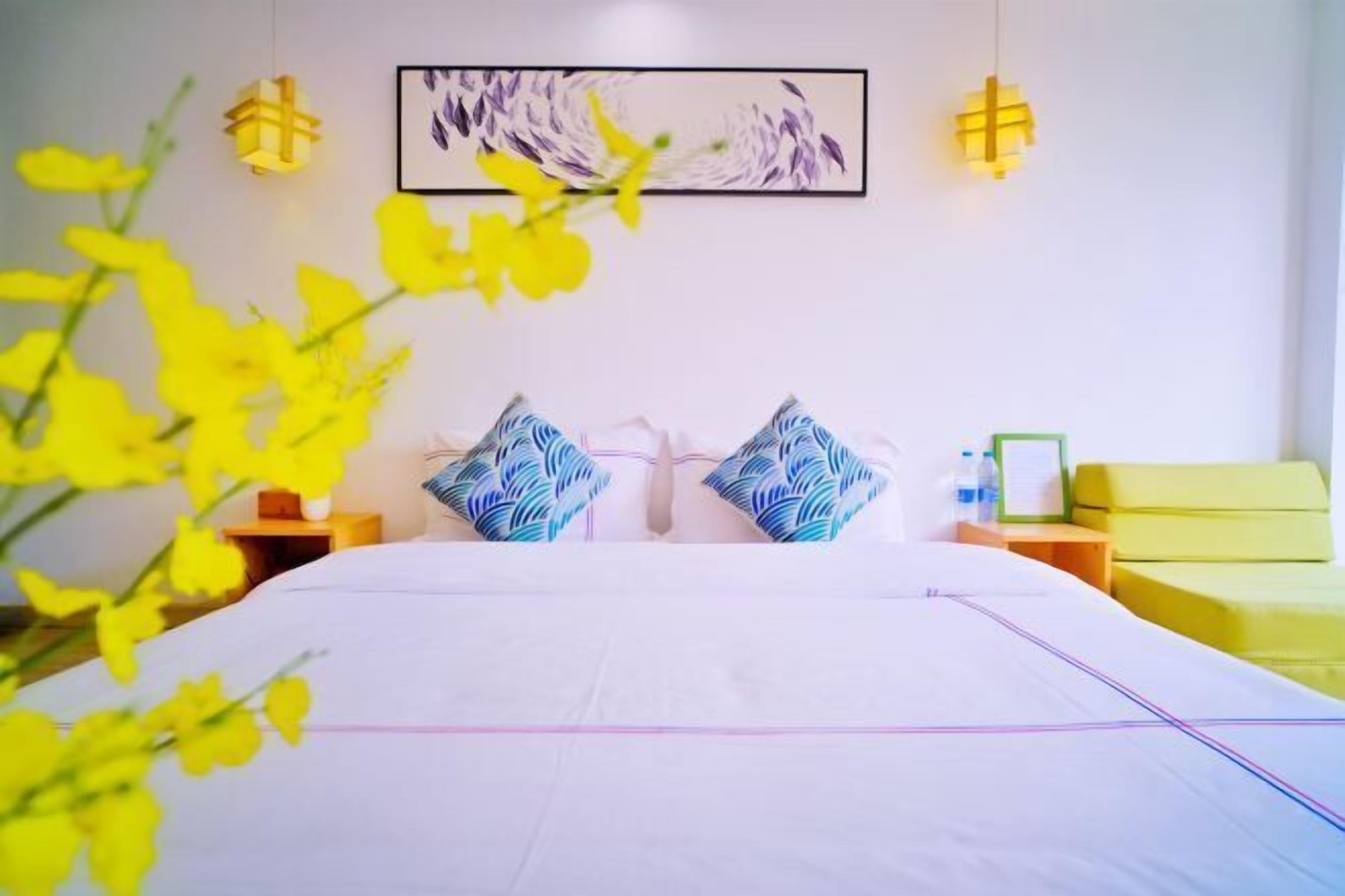 Habitación de hotel con una cama grande, dos almohadas, una mesita de noche y un sofá amarillo, además de una obra de arte colgada en la pared.