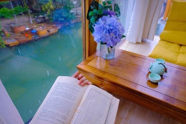 Una persona leyendo un libro junto a una ventana con vista a un cuerpo de agua y una silla amarilla.