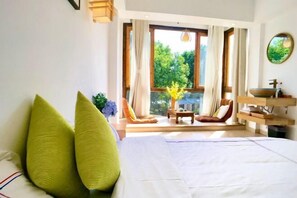 Un dormitorio con cama, dos almohadas, ventana con cortinas, una silla, una mesita y un baño con lavabo y espejo.