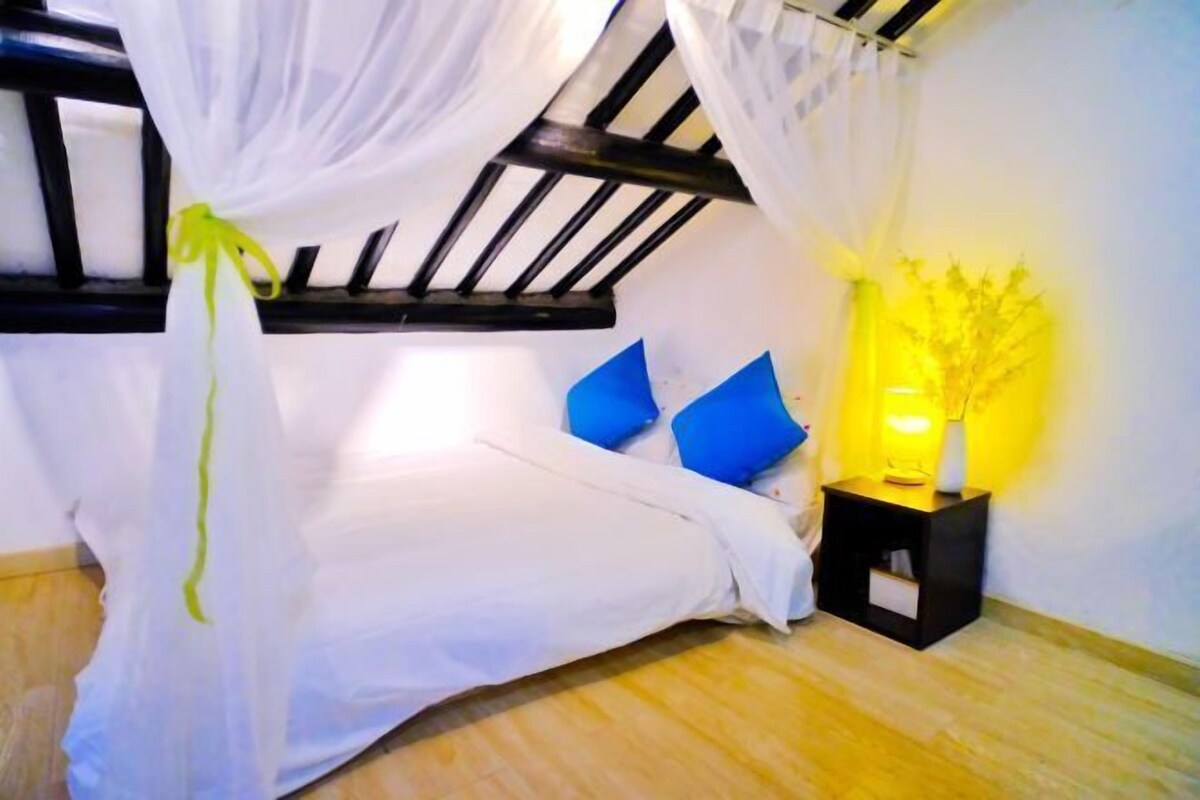 Un dormitorio con cama, cortinas blancas, almohadas azules, una mesita de noche con lámpara y suelo de madera.