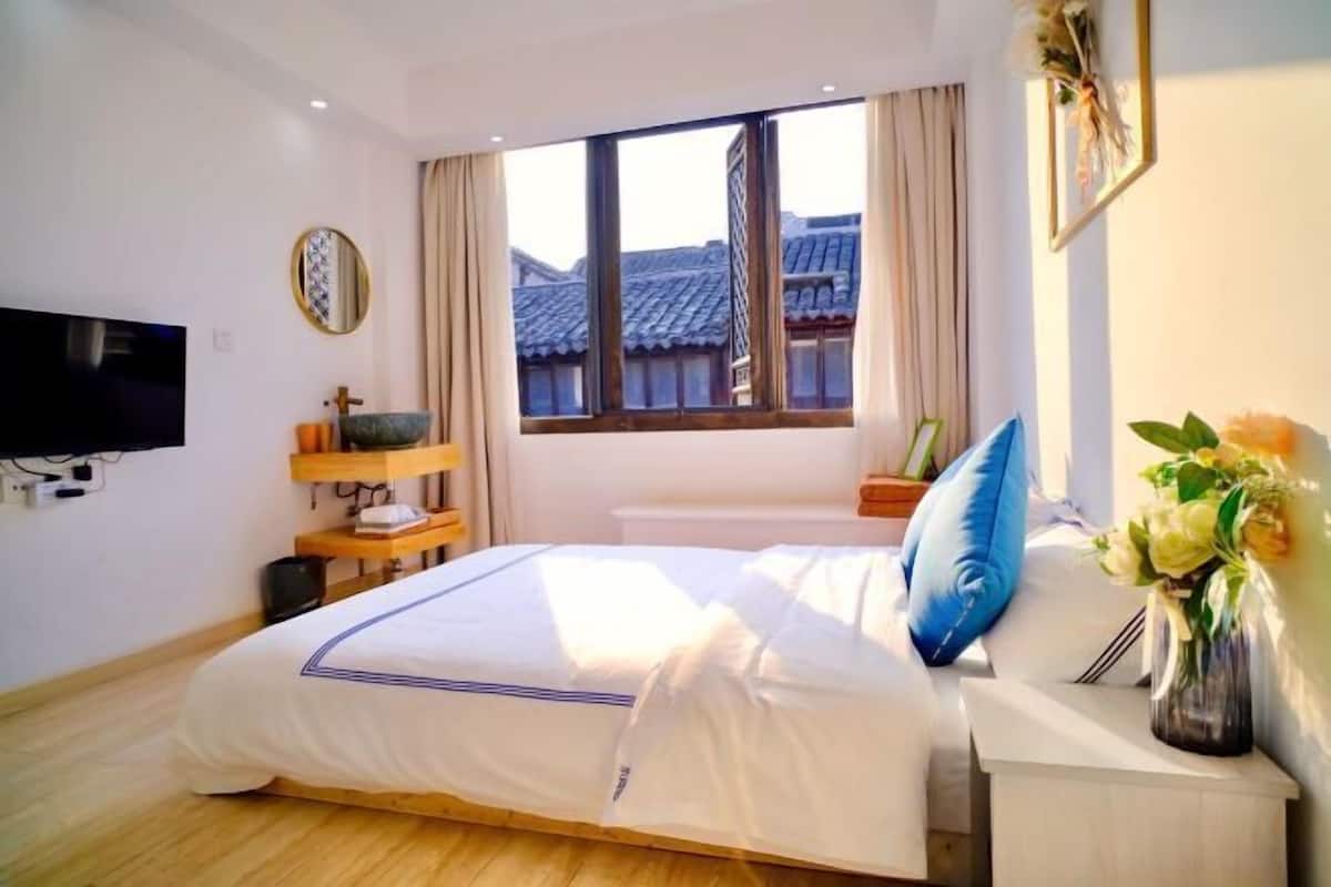 Une chambre à coucher comprenant un lit, une télévision, une fenêtre donnant sur un paysage où l’on peut admirer l’architecture traditionnelle, une table de chevet ornée d’un vase de fleurs et une horloge murale.