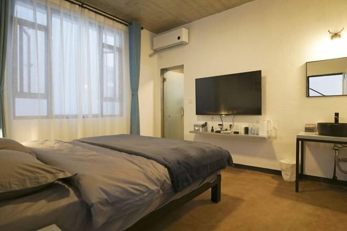 A bedroom with a bed, a TV mounted on the wall, a small table with a sink, and a window with sheer curtains.