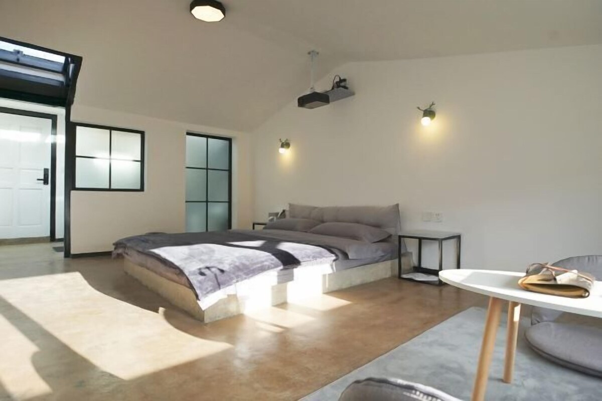 A modern bedroom with a bed, bedside tables, a small table, and a view of the outside.