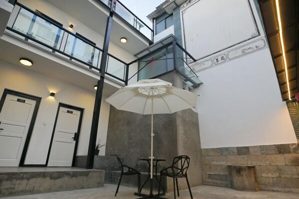A courtyard with a white umbrella, two chairs, and a table. There are doors and a staircase leading to an upper level.