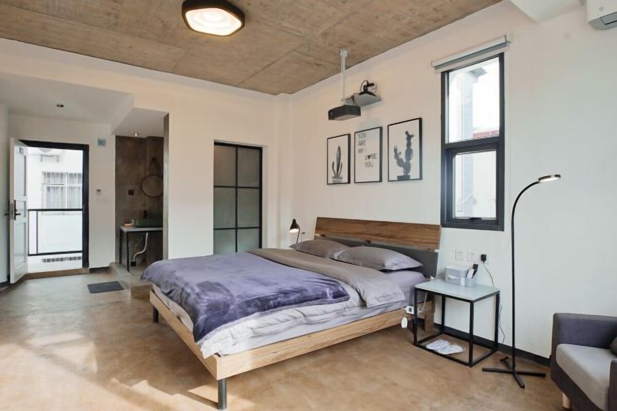 A modern bedroom with a wooden bed, a desk, and a chair. There are framed pictures on the wall and a window with blinds.